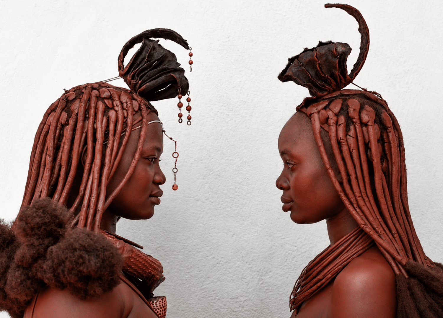Two Himba women in profile, wearing traditional ochre-covered hair and adornments